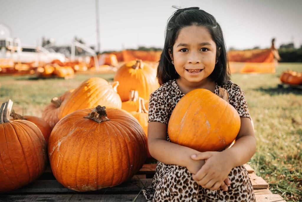 A young girl in a leopard-print dress sits outdoors holding a pumpkin, surrounded by several other pumpkins on a sunny day.