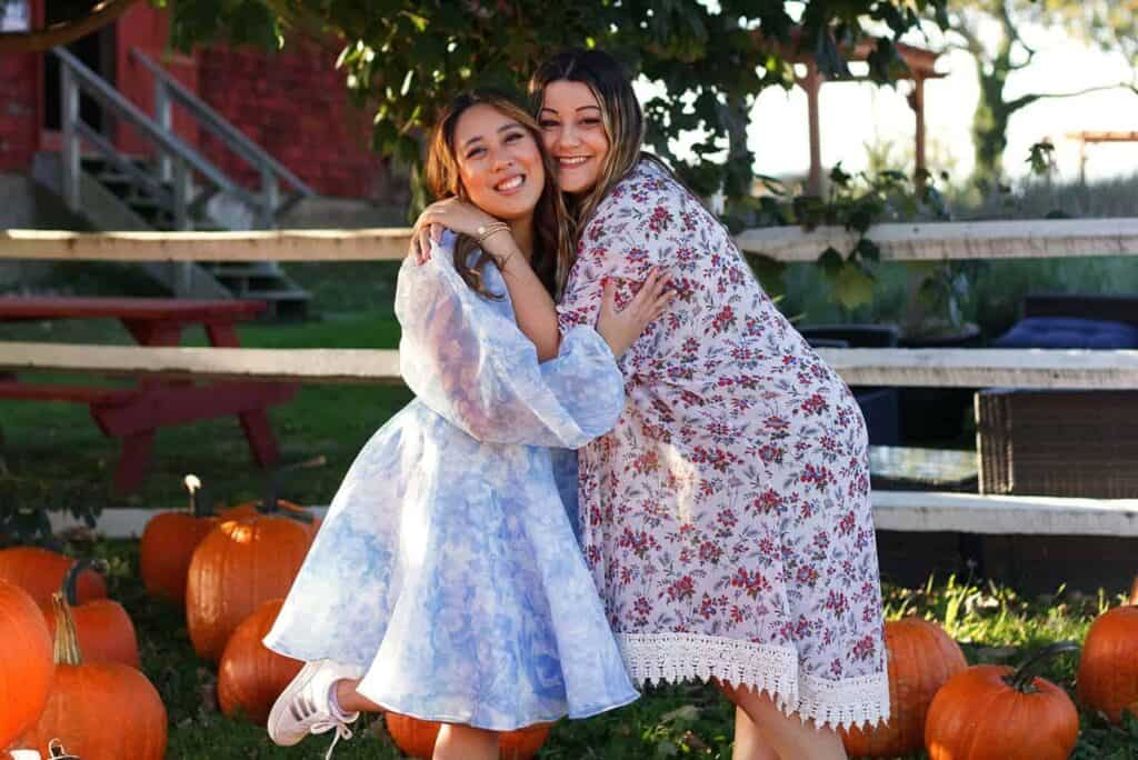 Two people, dressed in patterned dresses, stand outdoors amid pumpkins, embracing and smiling at the camera. A wooden fence and a red building are visible in the background.