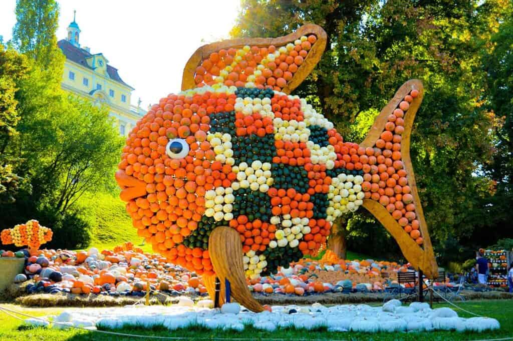 A large fish sculpture made from various colored pumpkins is displayed outdoors, set against a backdrop of trees and a multi-storied building.